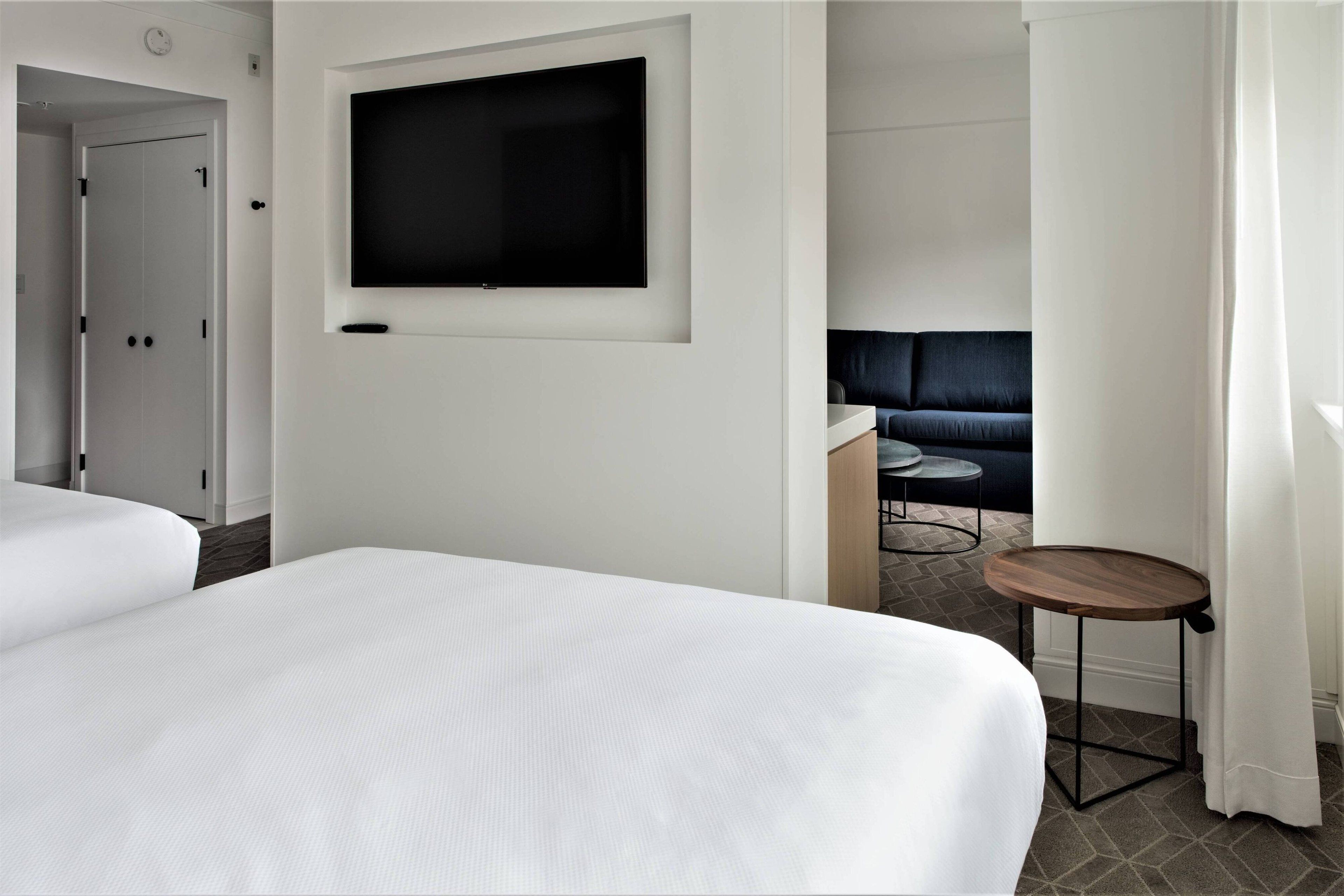 A hotel room with a large bed, a TV mounted on the wall, a small round table, and a dark-colored sofa in the background.