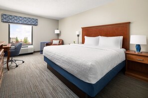 Una habitación de hotel con una cama grande, un escritorio, una silla, un ventanal y una mesita de noche con una lámpara.