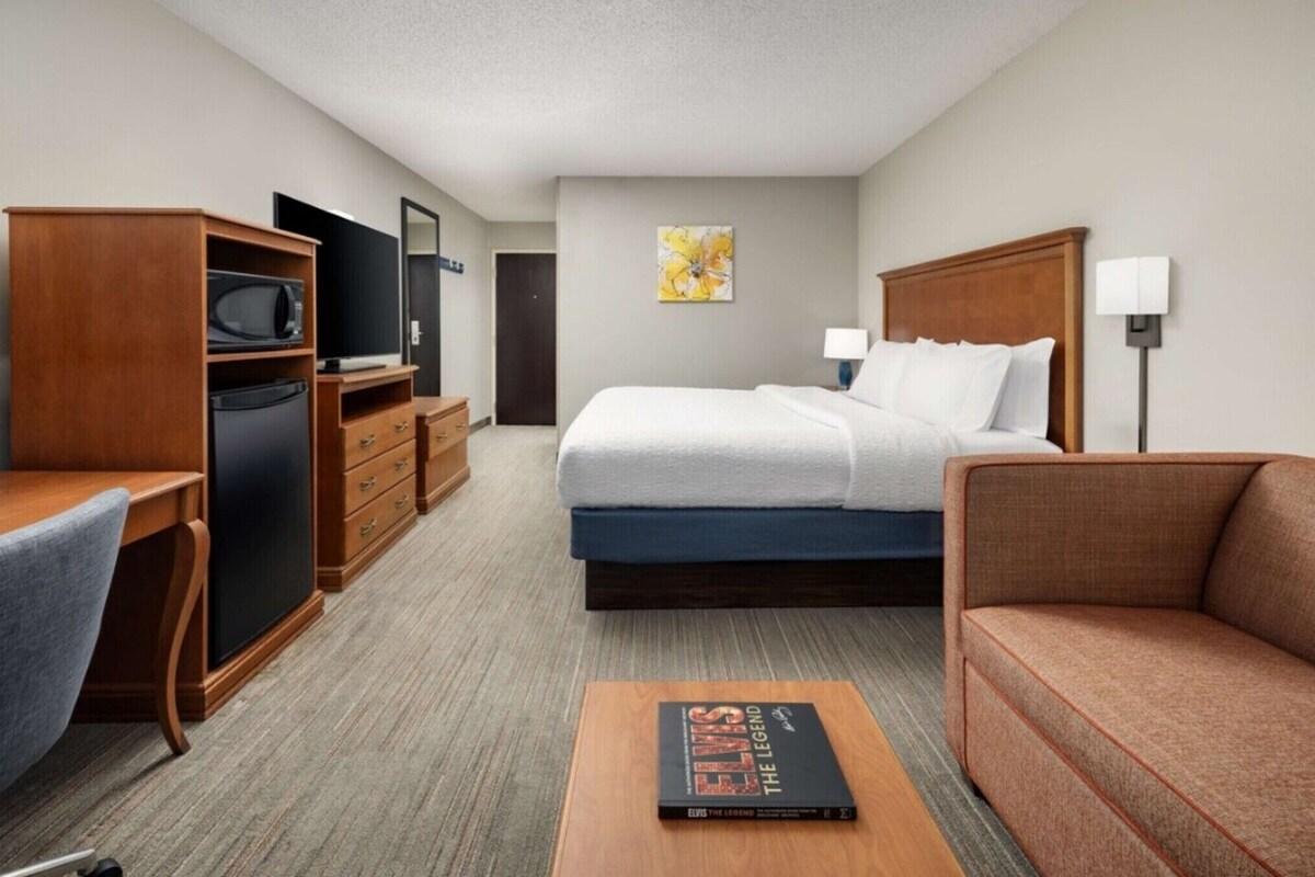 A hotel room with a bed, a desk, a chair, a sofa, a TV, a microwave, a dresser, and a book on the table.