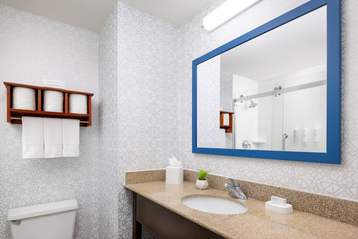 Un baño moderno con encimera de granito, un lavamanos blanco, un espejo grande con marco azul y un perchero con toallas blancas.