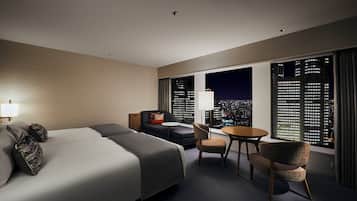 Habitación de hotel con una cama grande, un sofá, una mesita y una silla. Dos ventanas ofrecen vistas a la ciudad.
