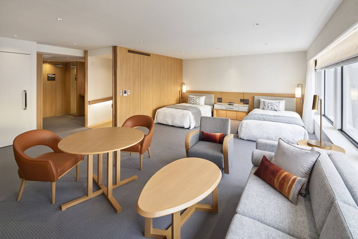 A modern hotel room with a sofa, two armchairs, a round table, a bed, and a small desk.