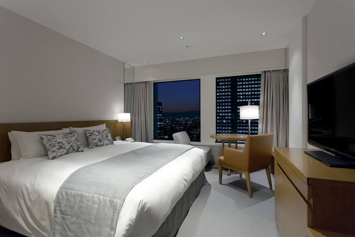 A hotel room with a large bed, a desk with a chair, a TV, and a window with a city view.