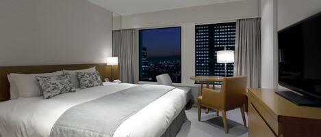 A hotel room with a large bed, a desk with a chair, a TV, and a window with a city view.