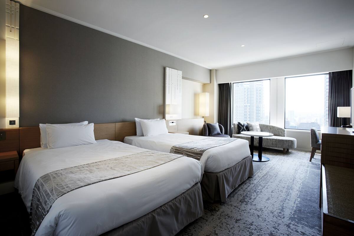 A hotel room with two beds, a sofa, a desk, and a large window offering a city view.