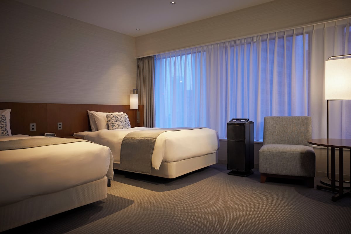A hotel room with two beds, a desk, a chair, and a lamp.