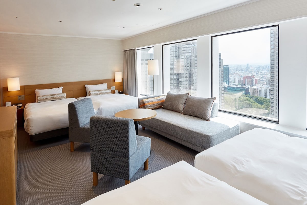 A hotel room with two beds, a sofa, a chair, and a small table. The room has large windows with a city view.
