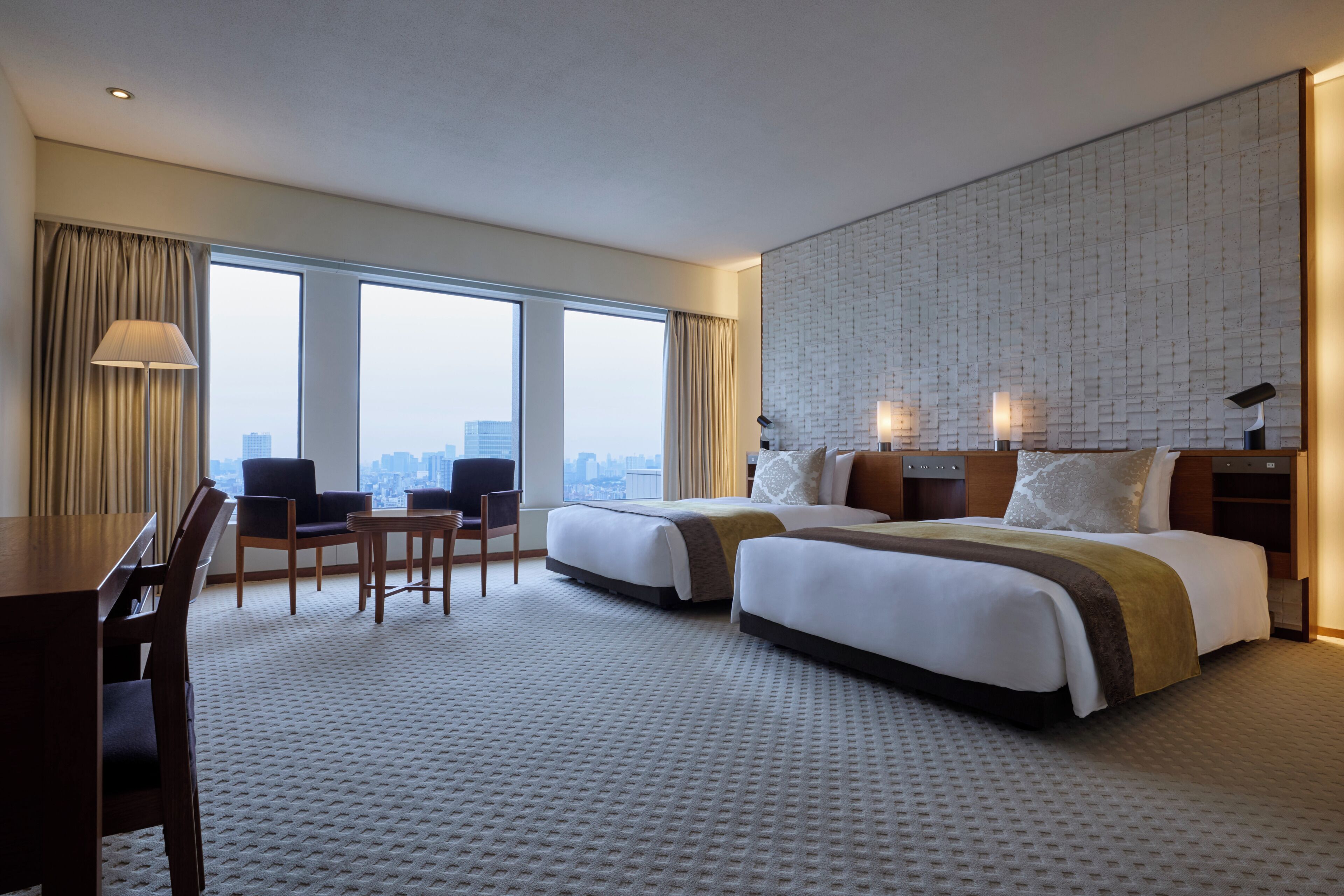 Habitación de hotel con dos camas, un escritorio, sillas, un ventanal con vistas a la ciudad y una pared con textura.
