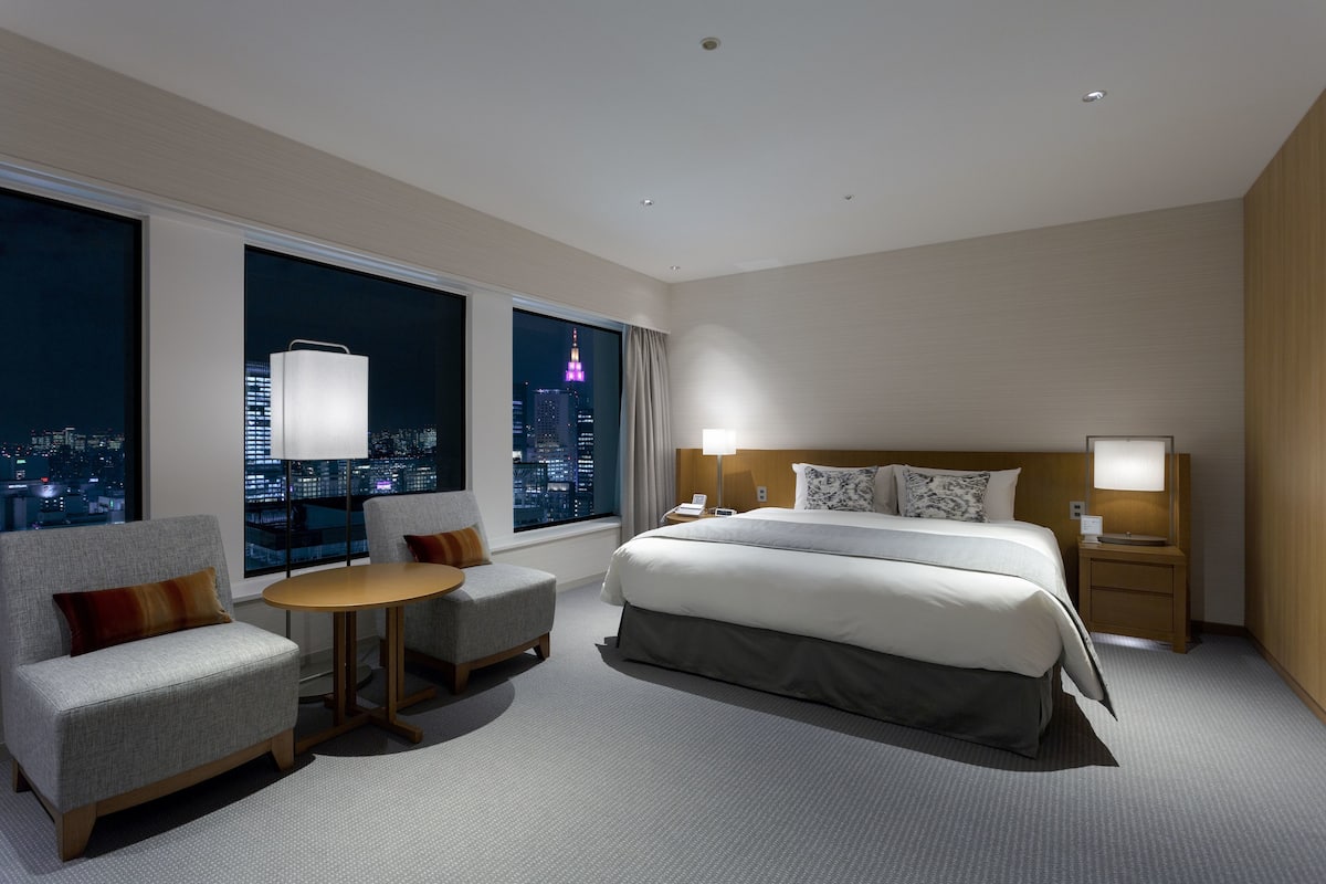 A modern hotel room with a large bed, a desk, a chair, and a view of a cityscape at night.