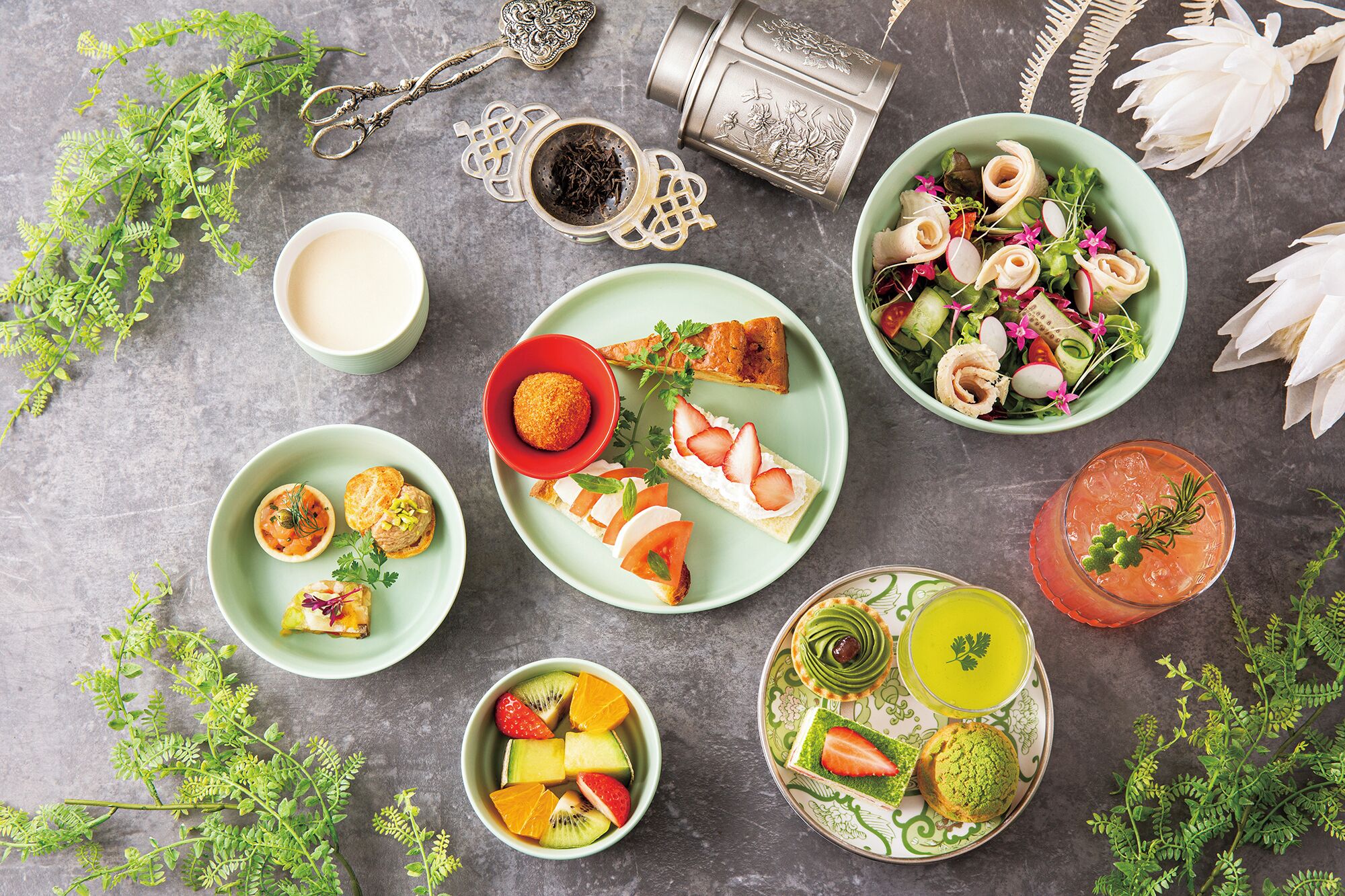 A variety of food items and drinks arranged on a table, including a salad, fruit, pastries, and a cocktail.