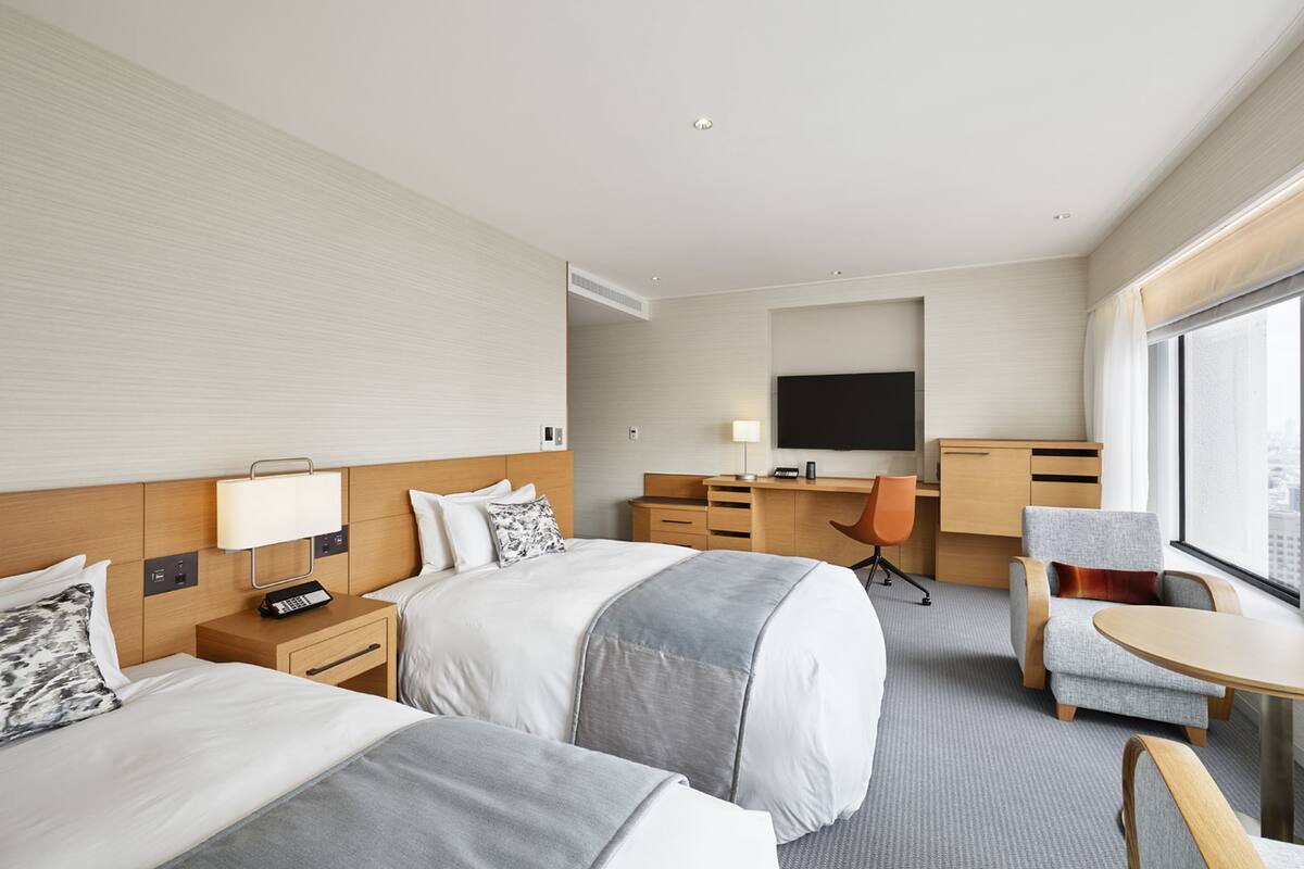 A hotel room with two beds, a desk, a chair, a TV, and a large window.