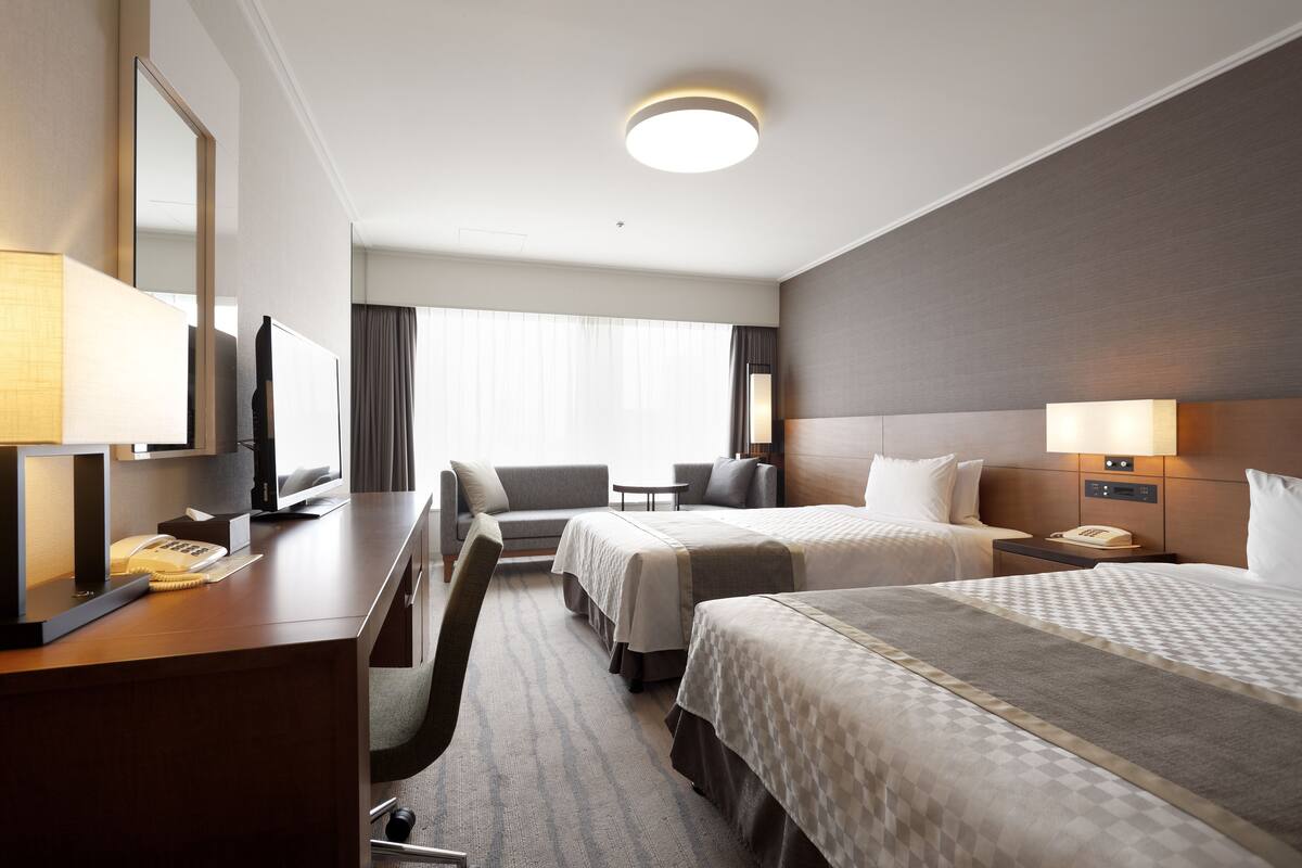 A hotel room with two beds, a desk with a telephone and a lamp, a chair, a bedside table with a lamp, and a large window with curtains.
