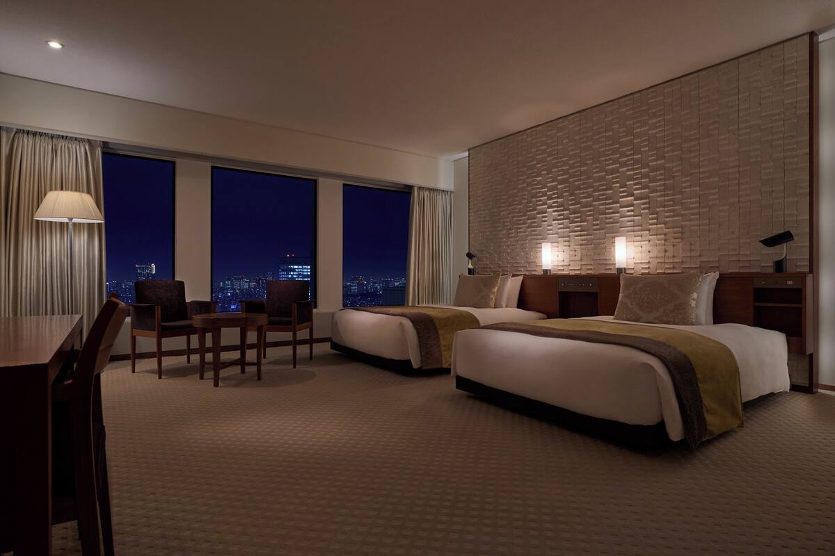 A hotel room with two beds, a desk, and a chair. There are large windows with a city view.