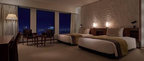 A hotel room with two beds, a desk, and a chair. There are large windows with a city view.