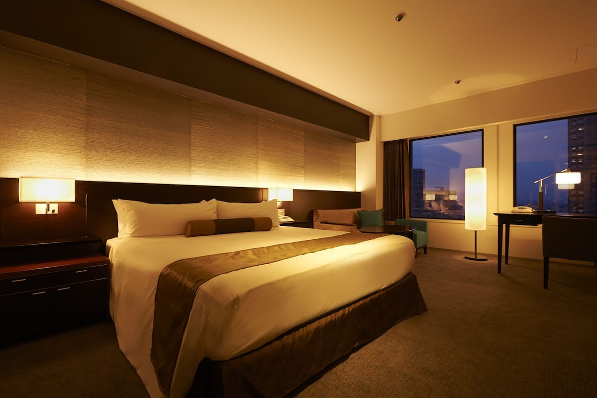 A hotel room with a large bed, a desk, a chair, and a view of the city at night.