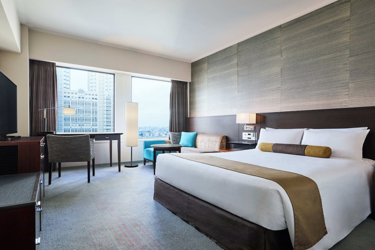 A hotel room with a large bed, a desk, a chair, a small table, and a view of a cityscape through large windows.