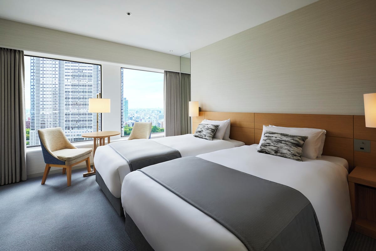 A hotel room with two beds, a chair, a small table, and a view of a cityscape through large windows.