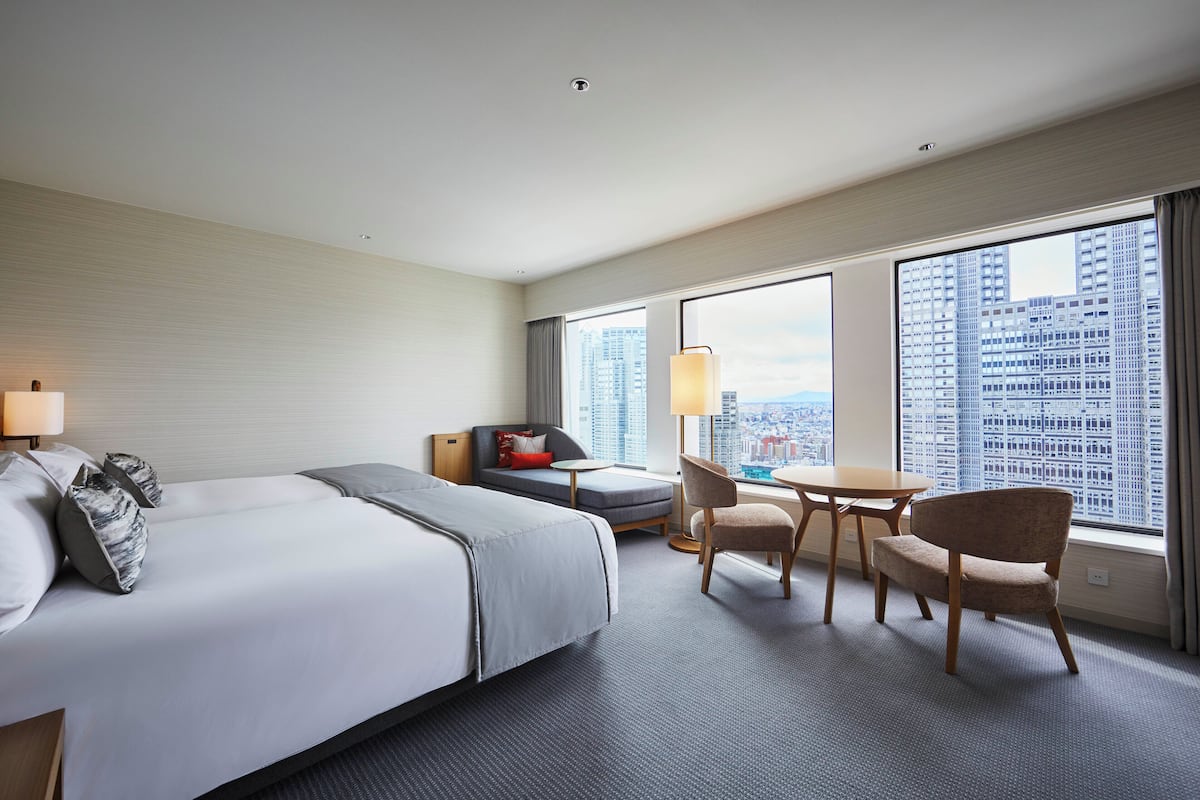 A hotel room with a large bed, a sofa, a dining table, and chairs. There is a city view through the windows.