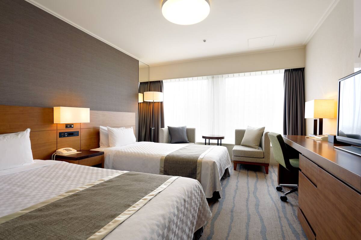 A hotel room with two beds, a desk, a chair, a TV, and a small table.