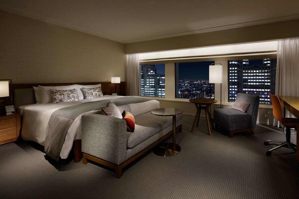 A hotel room with a bed, a sofa, a desk, and a chair. There is a window with a city view.