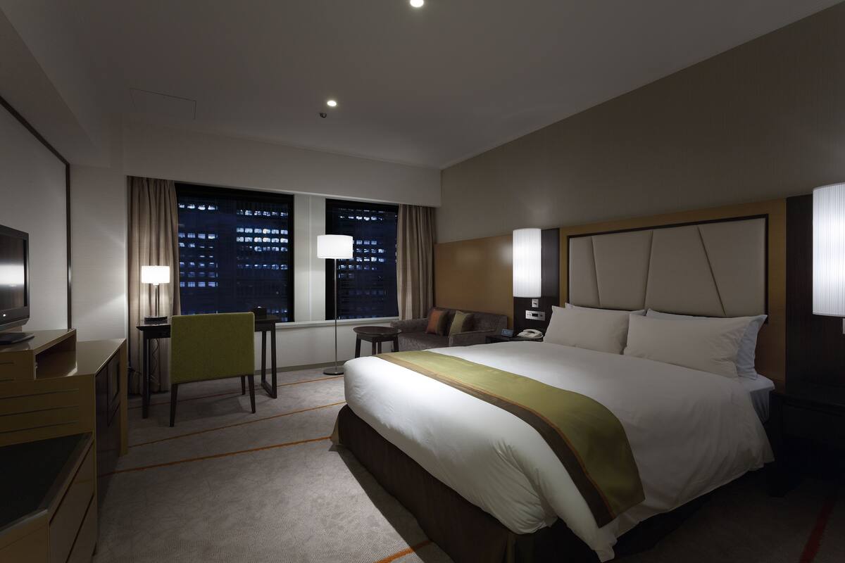 A hotel room with a large bed, a desk, a chair, a TV, and a window with a city view.