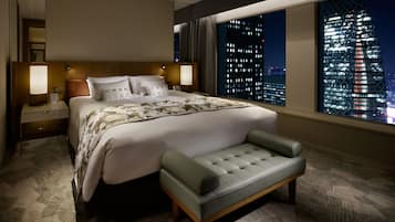 A hotel room with a large bed, a bench, a nightstand, a lamp, and a view of a cityscape at night.