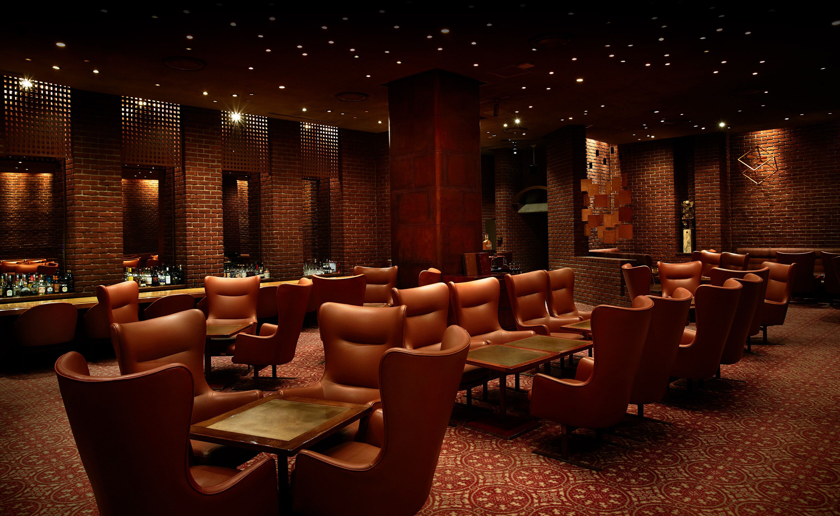 Ein gedämpft beleuchteter Lounge-Bereich mit Ledersesseln und Tischen, Backsteinwänden und einer Bar im Hintergrund.