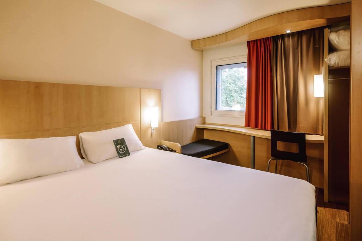 A hotel room with a large bed, a desk, a chair, a window with curtains, and a bedside table.