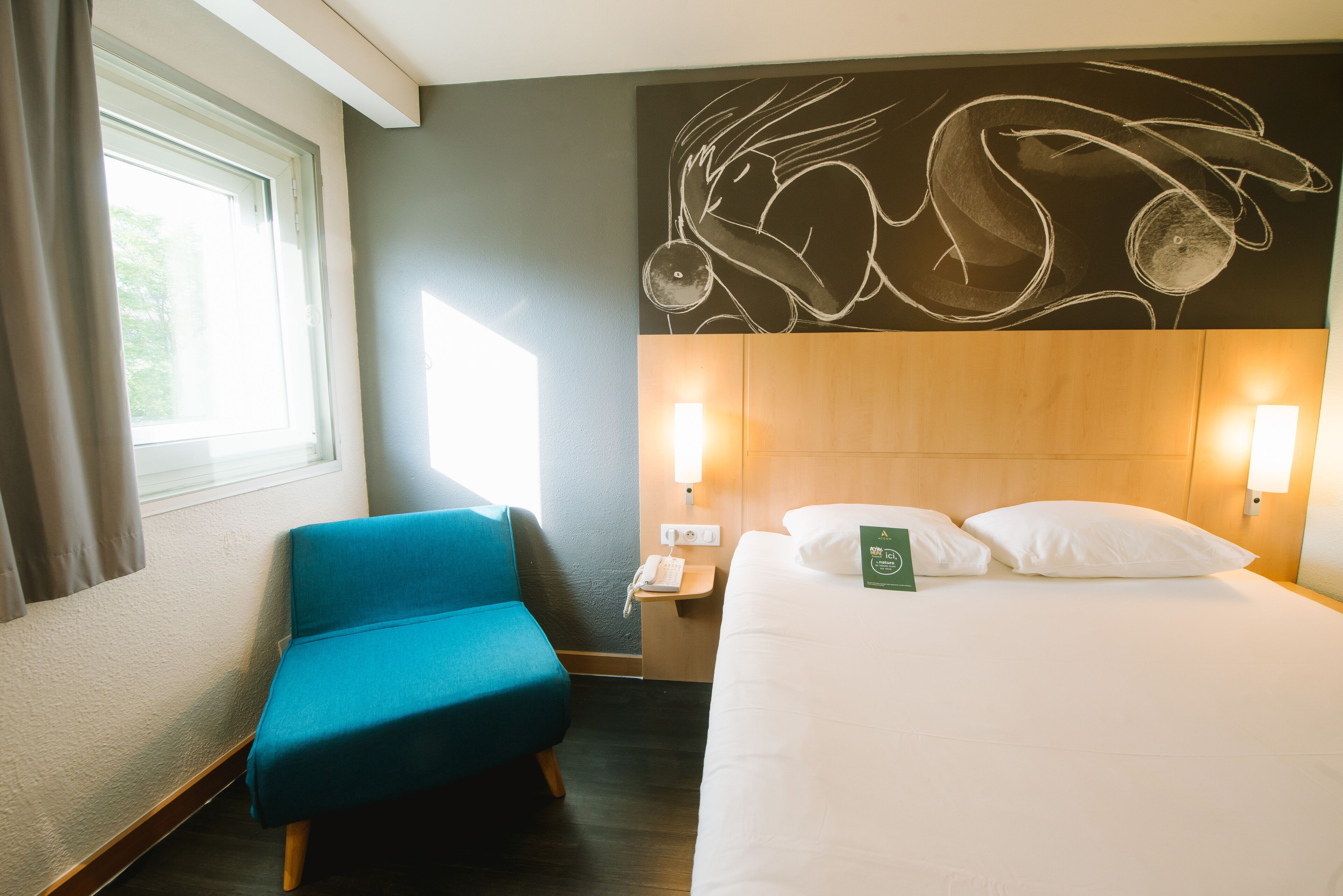A hotel room with a large bed, a blue armchair, a wall art piece, and a window with curtains.