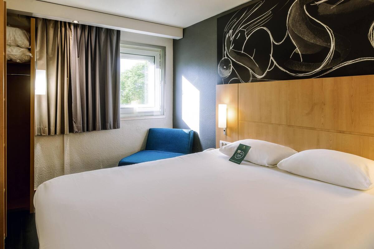 A hotel room with a large bed, a blue chair, a window with curtains, and a wall art piece.