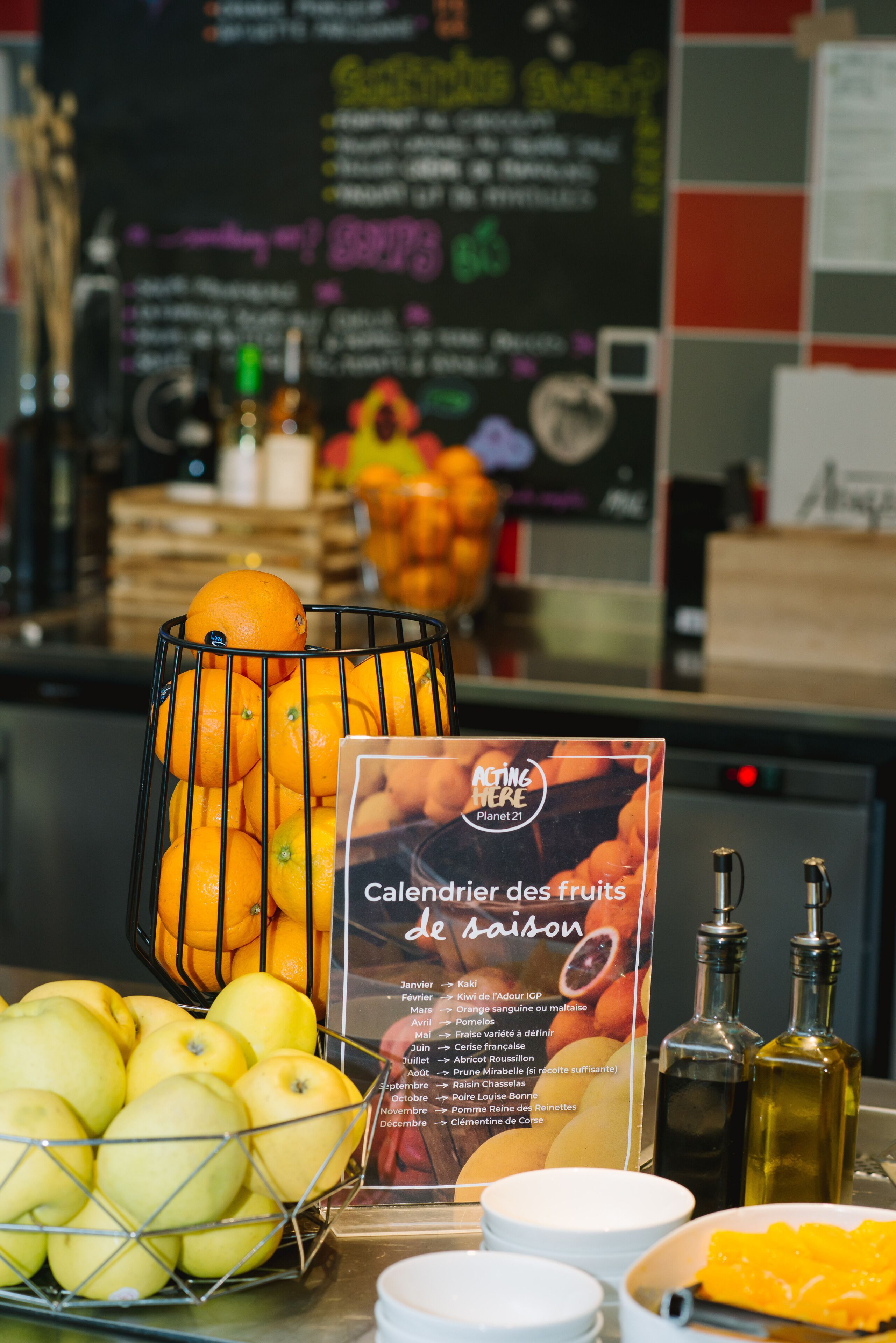 A fruit display with oranges and apples, a calendar of seasonal fruits, and a menu board in the background.
