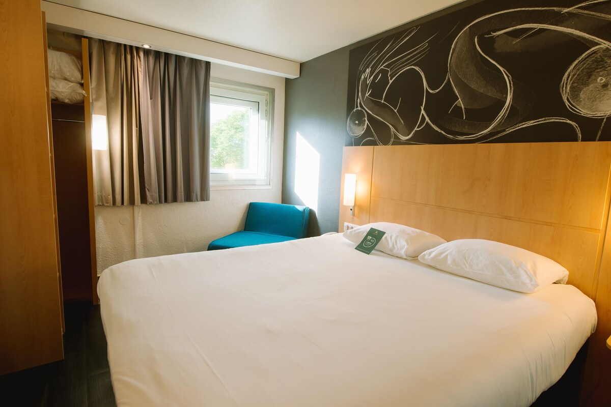 A hotel room with a large bed, a blue chair, a wardrobe, and a window with curtains.