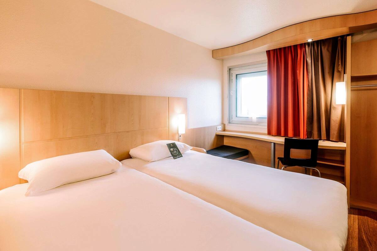 A hotel room with two beds, a desk, a chair, a window with curtains, and a wall-mounted light.