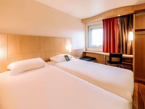 Ett hotellrum med två sängar, ett skrivbord, en stol, ett fönster med gardiner och en väggmonterad lampa.
