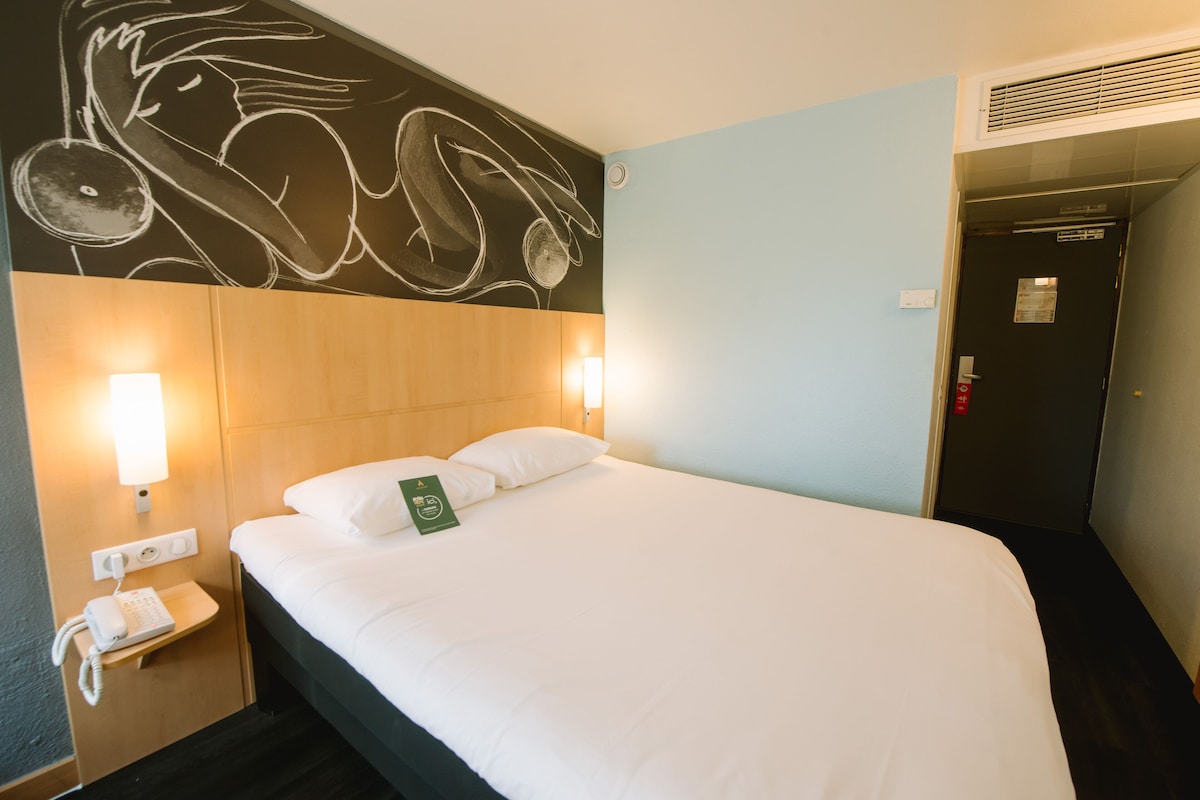 A hotel room with a bed, a wall-mounted phone, and a door with a red emergency button.