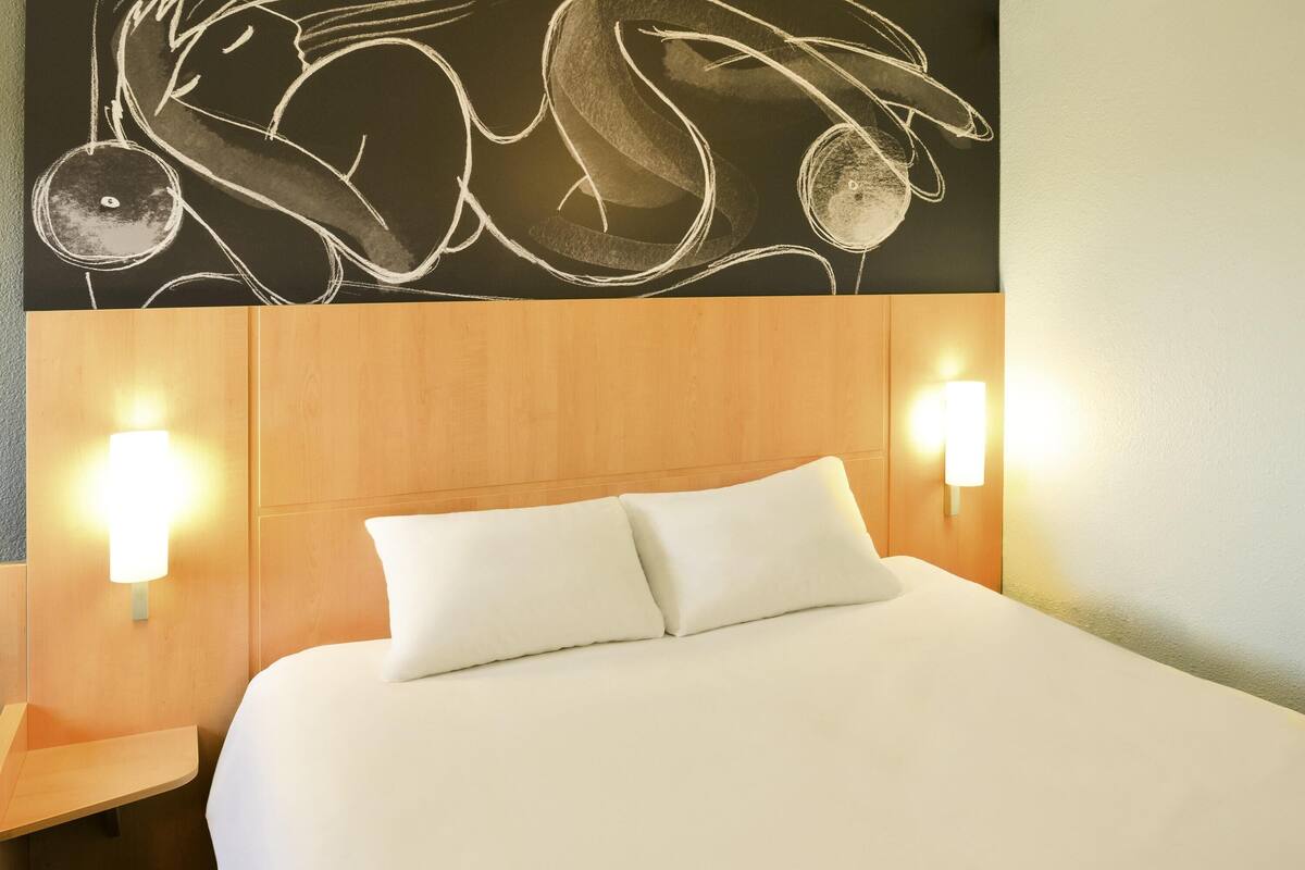 A hotel room with a wooden headboard, two white pillows, and wall-mounted light fixtures.