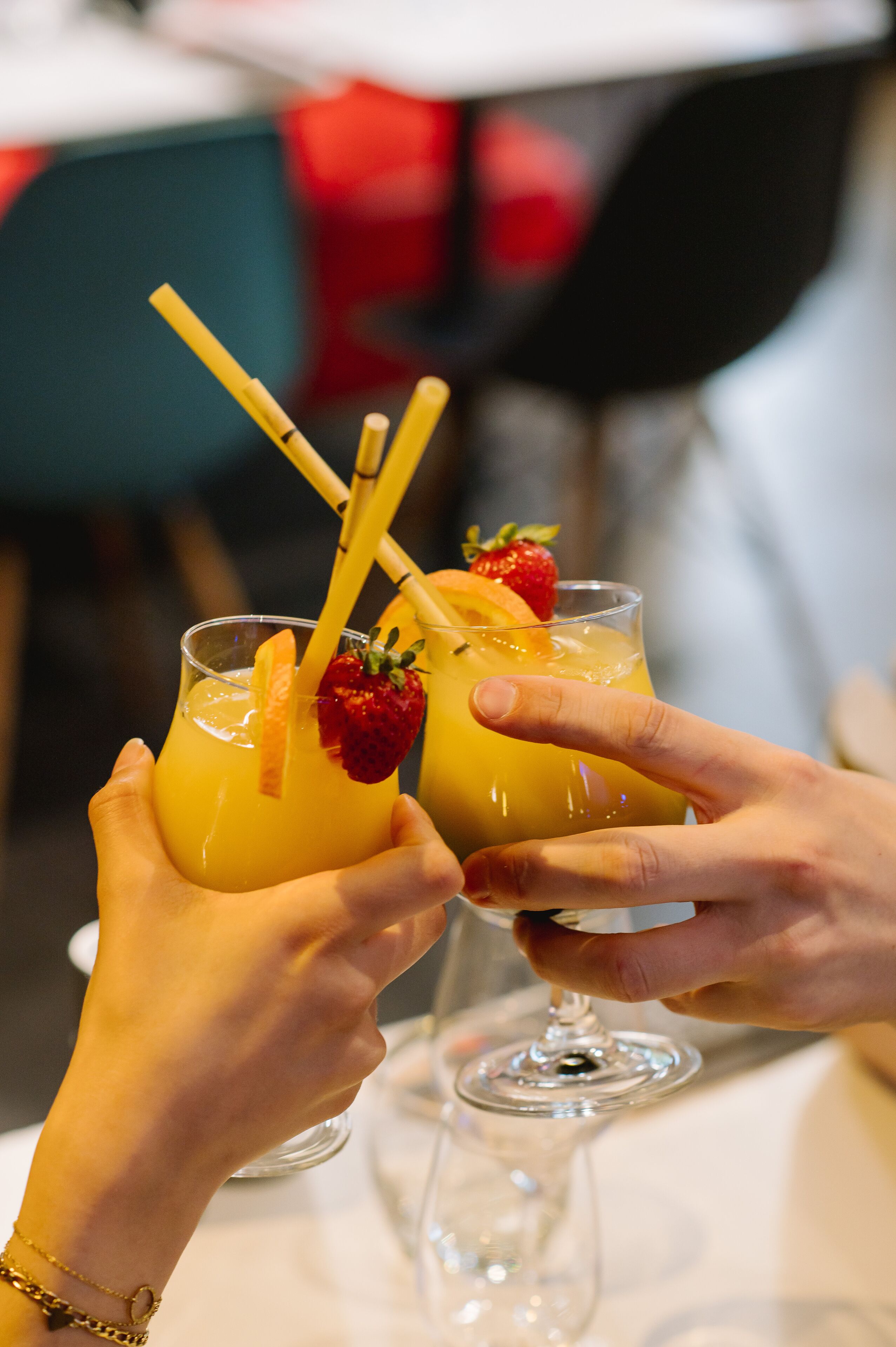 Two hands holding glasses of a yellow beverage with a strawberry and orange garnish.