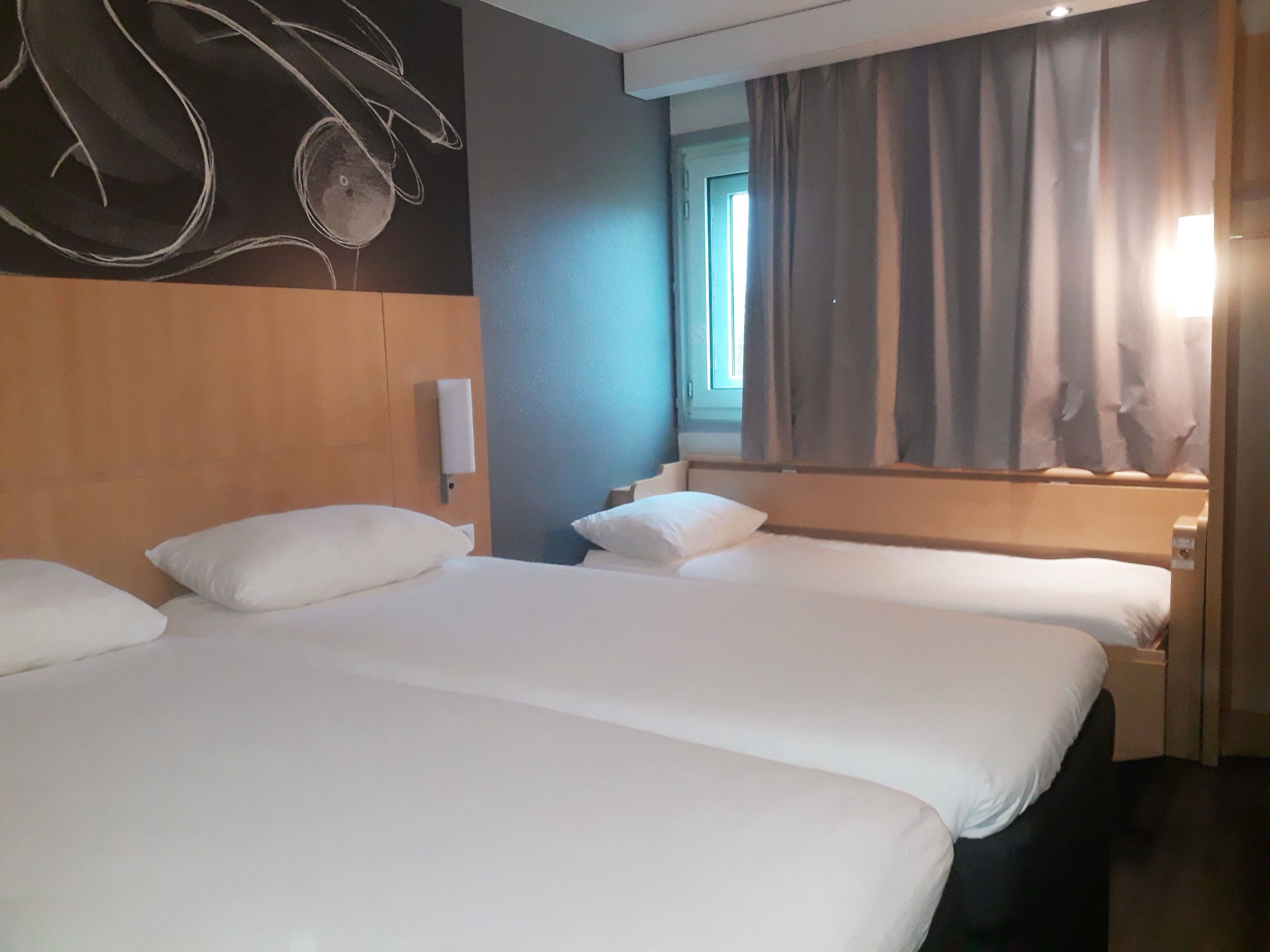 A hotel room with two beds, a wooden headboard, a wall-mounted light, and a window with curtains.
