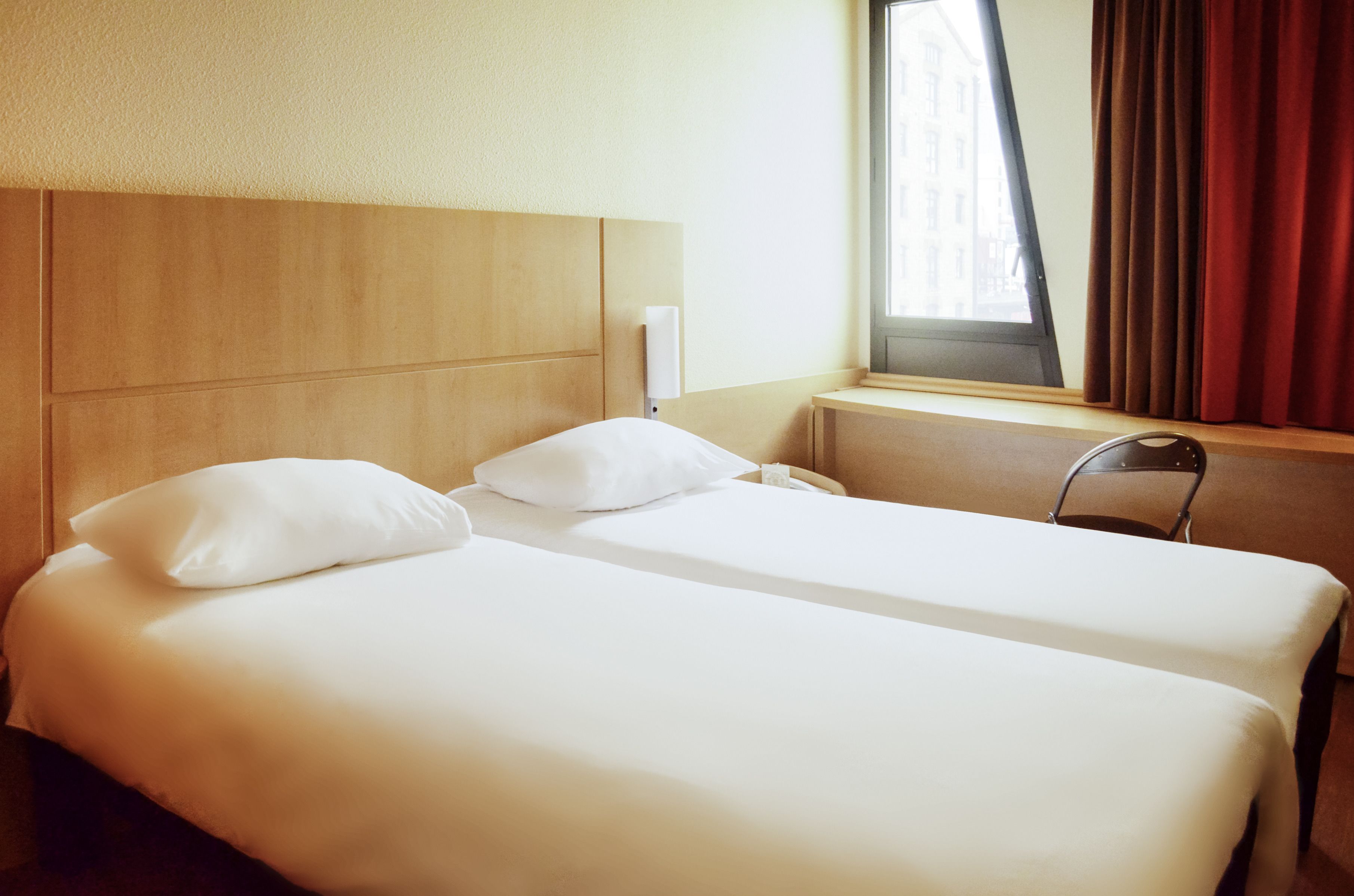 A hotel room with two single beds, a wooden headboard, a window with curtains, and a chair.