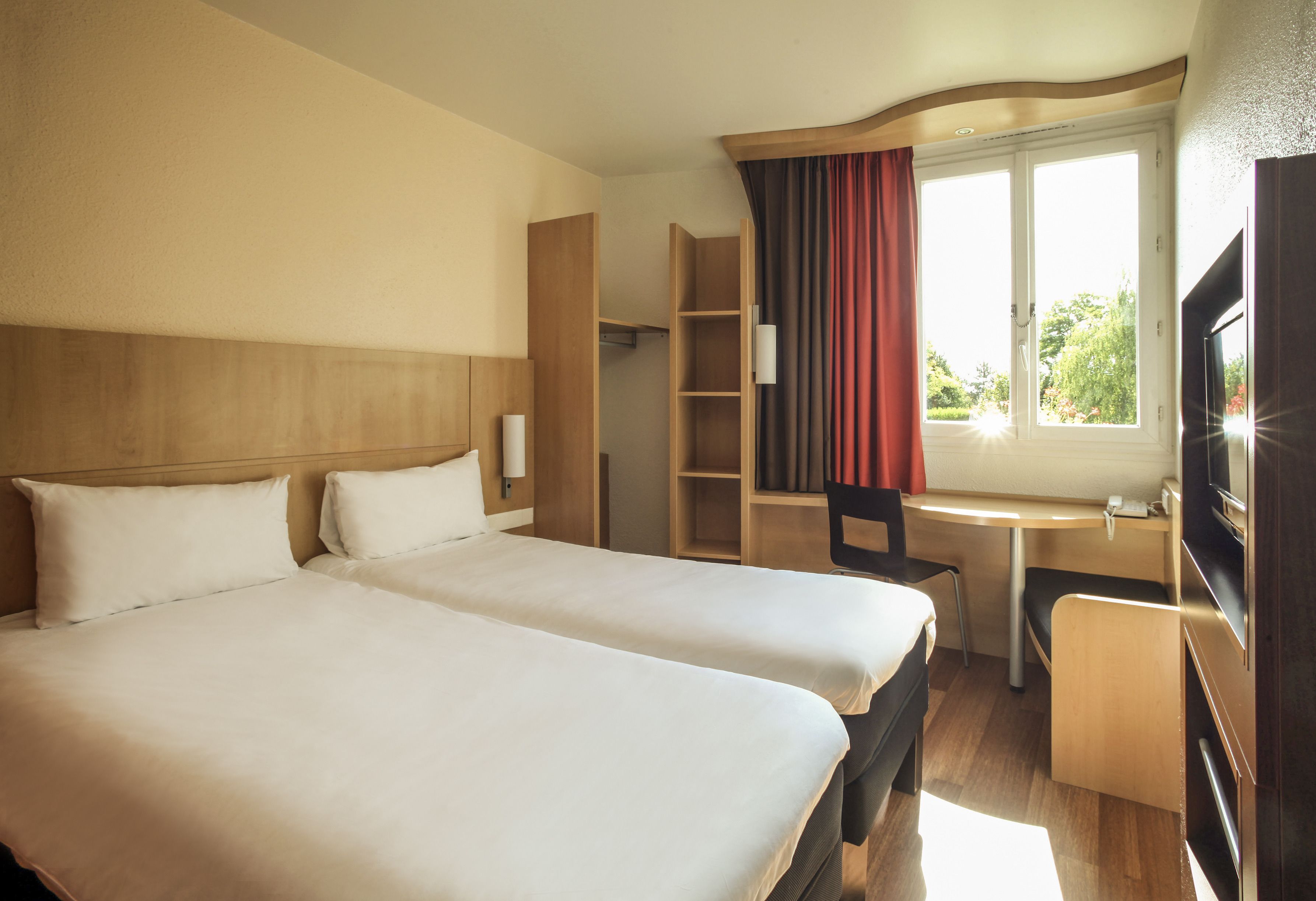 A hotel room with two beds, a desk, a chair, a television, and a window with curtains.