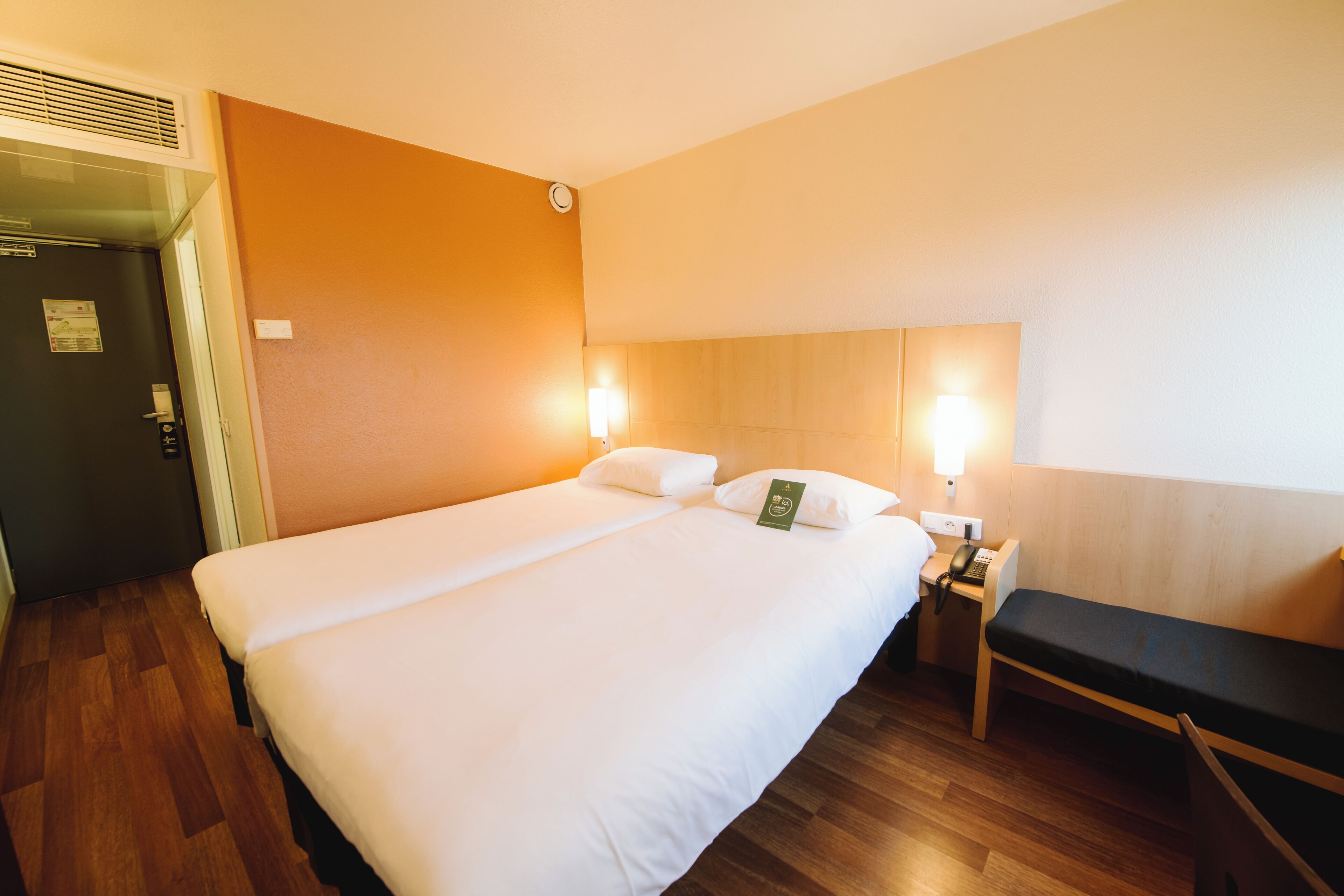 Hotelkamer met twee bedden, een houten vloer, een bankje en een nachtkastje met telefoon.