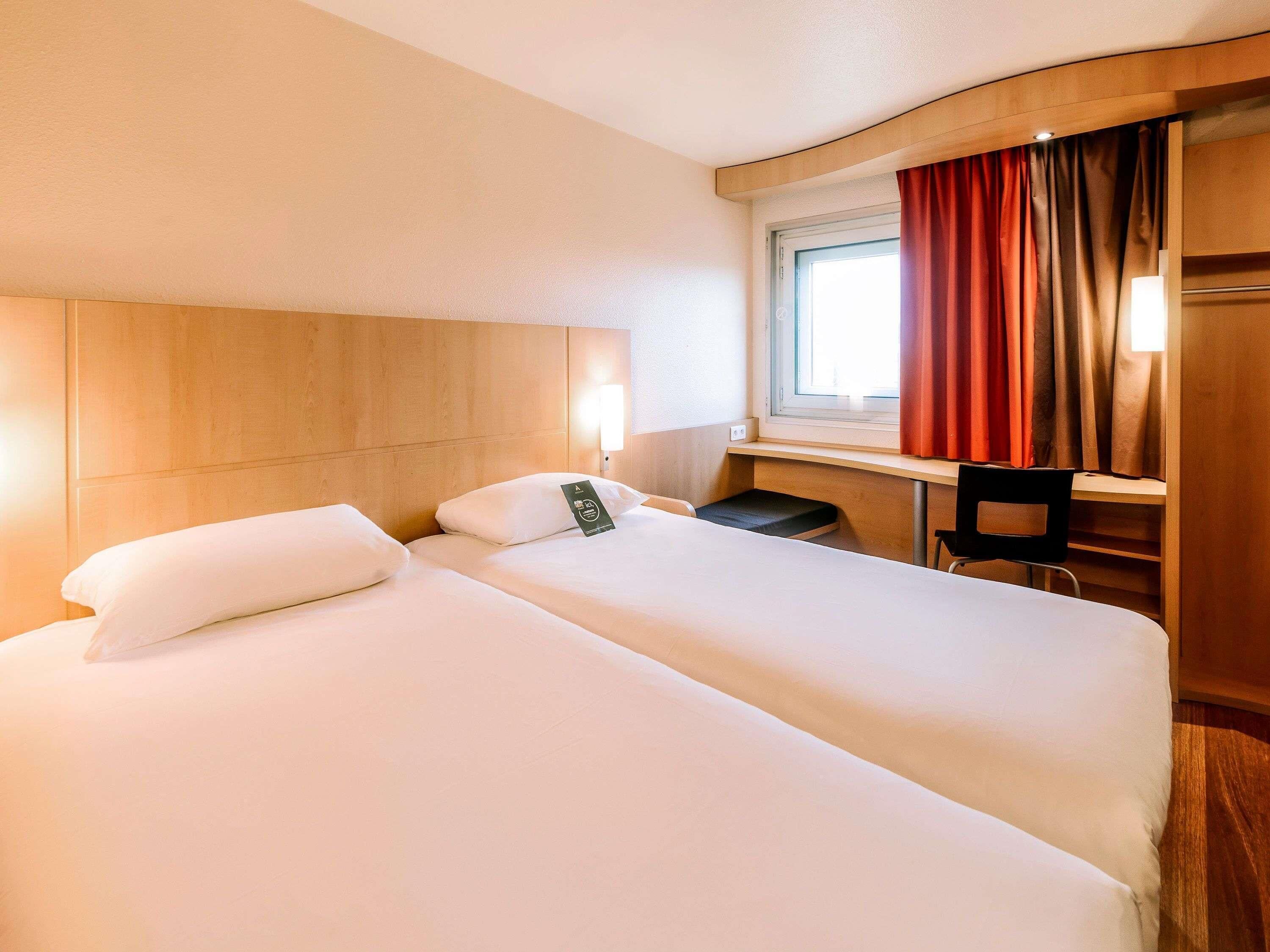 Camera d'albergo con due letti, una scrivania, una sedia, una finestra con tende e una lampada a parete.