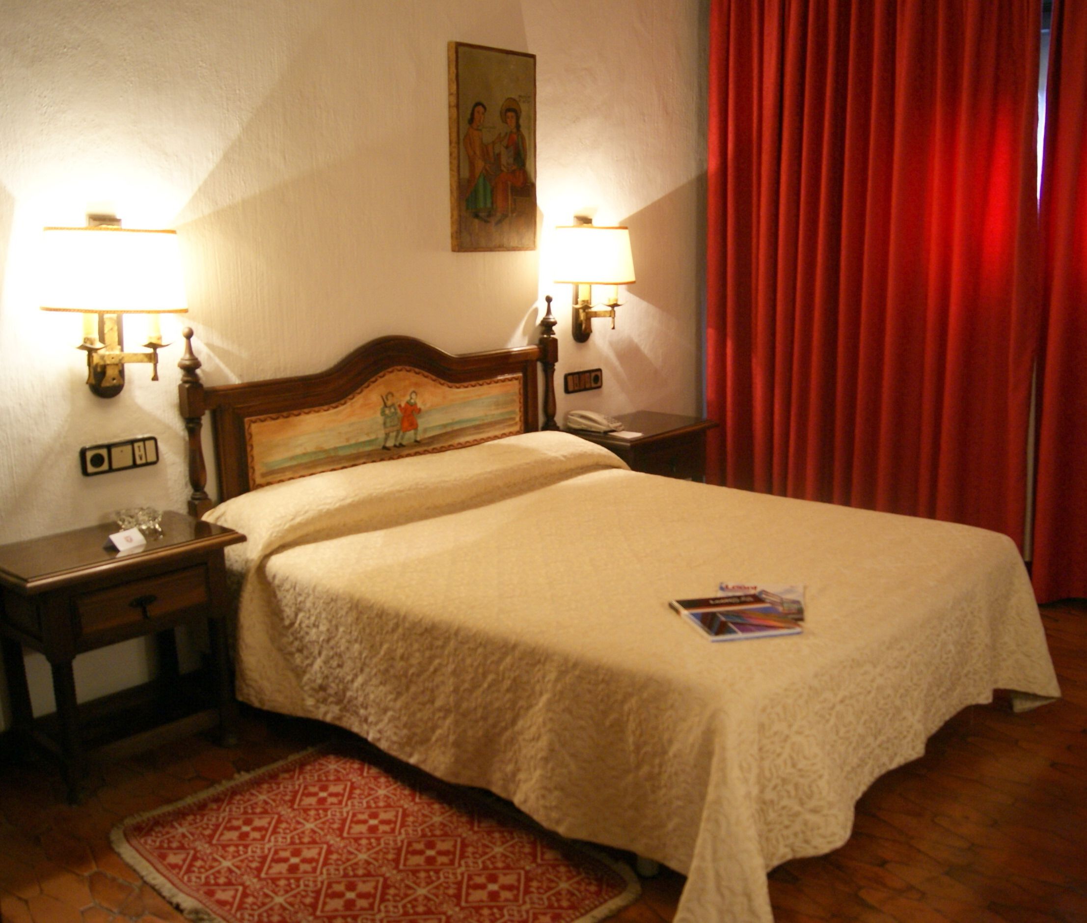 Habitación de hotel con una cama grande, dos mesitas de noche con lámparas, una cortina roja y un cuadro en la pared.