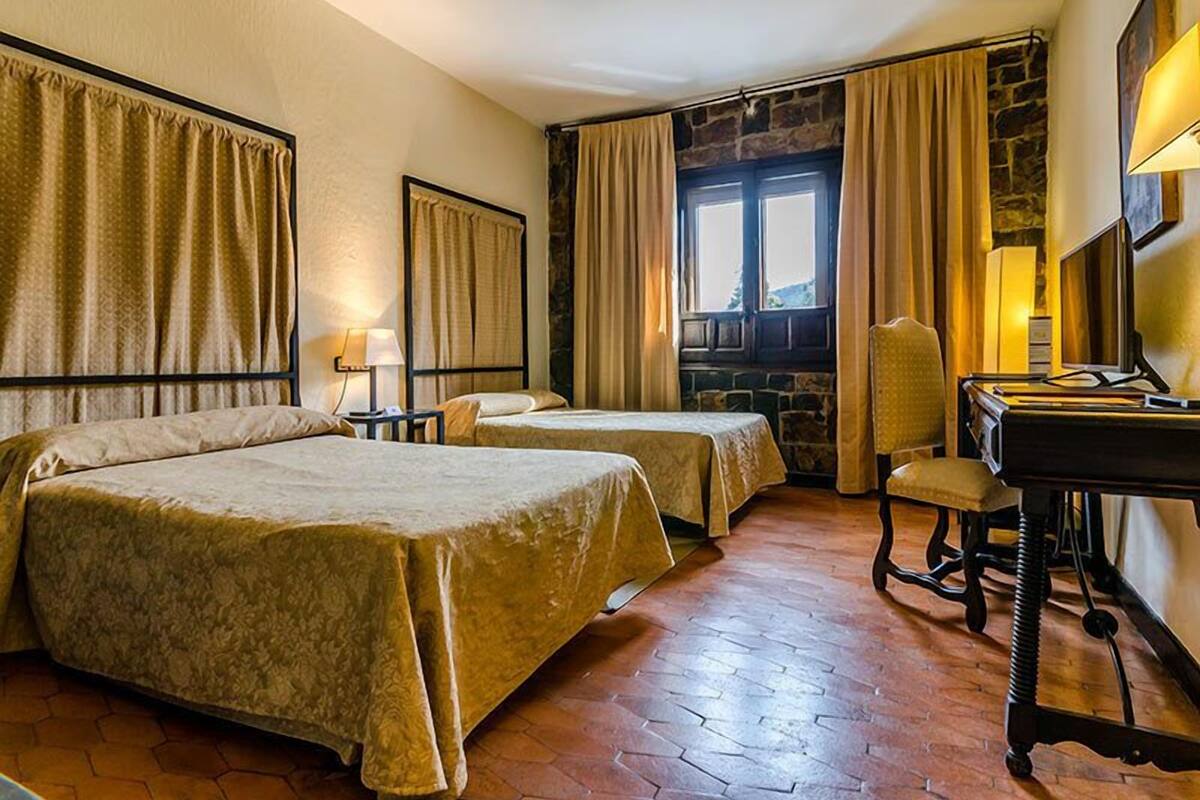 A hotel room with two beds, a desk with a chair, a stone wall, and a window with curtains.
