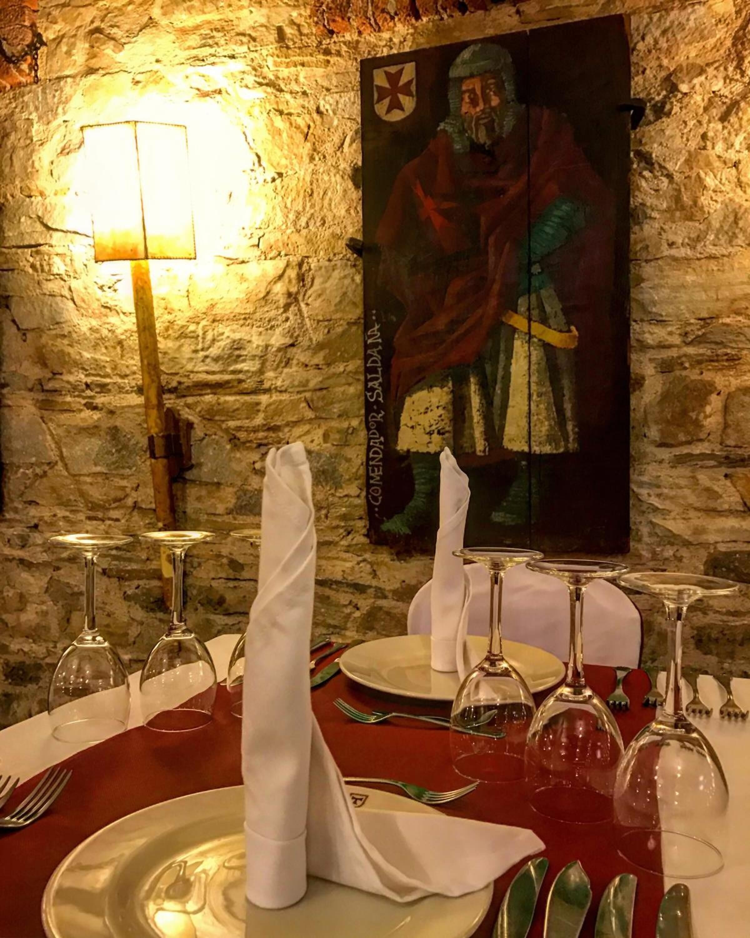 Mesa lista para comer, con copas de vino, platos y cubiertos. Se observa un cuadro en la pared detrás de la mesa.