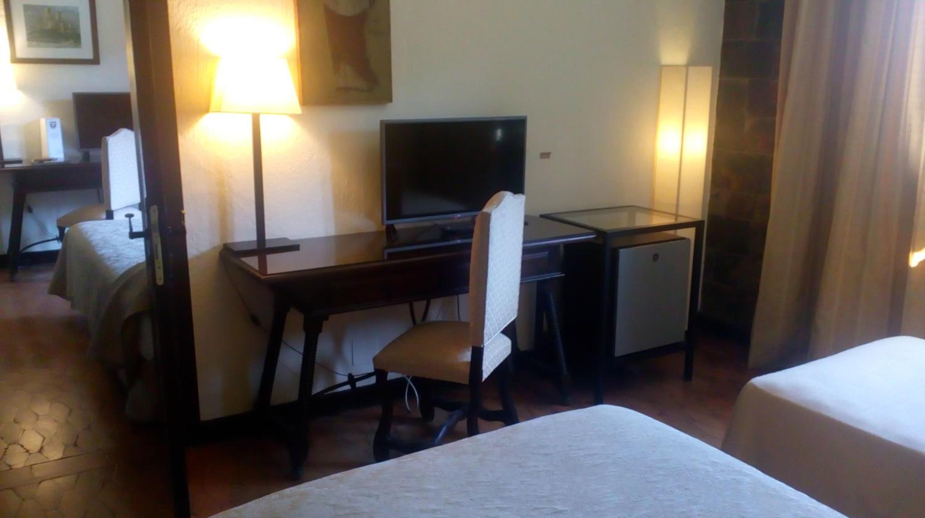 Una habitación de hotel con una cama individual, un escritorio con computadora, un refrigerador pequeño y una lámpara.