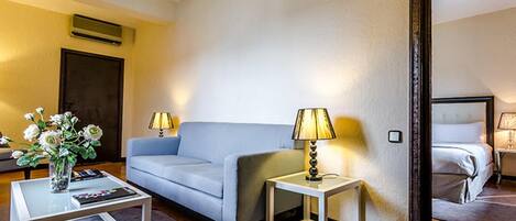 A hotel room with a light blue sofa, a glass coffee table, a side table with a lamp, and a bed in the adjacent room.