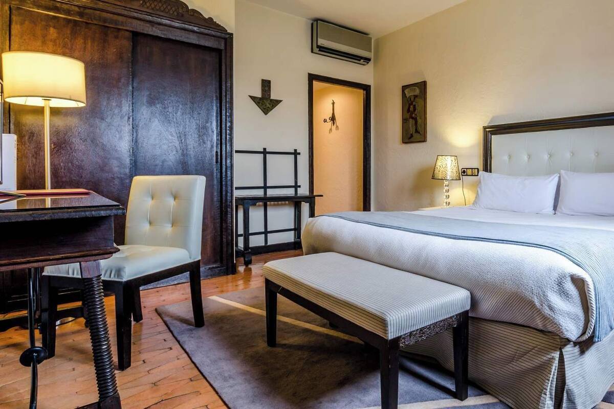 A hotel room with a large bed, a desk, a chair, and a bench.