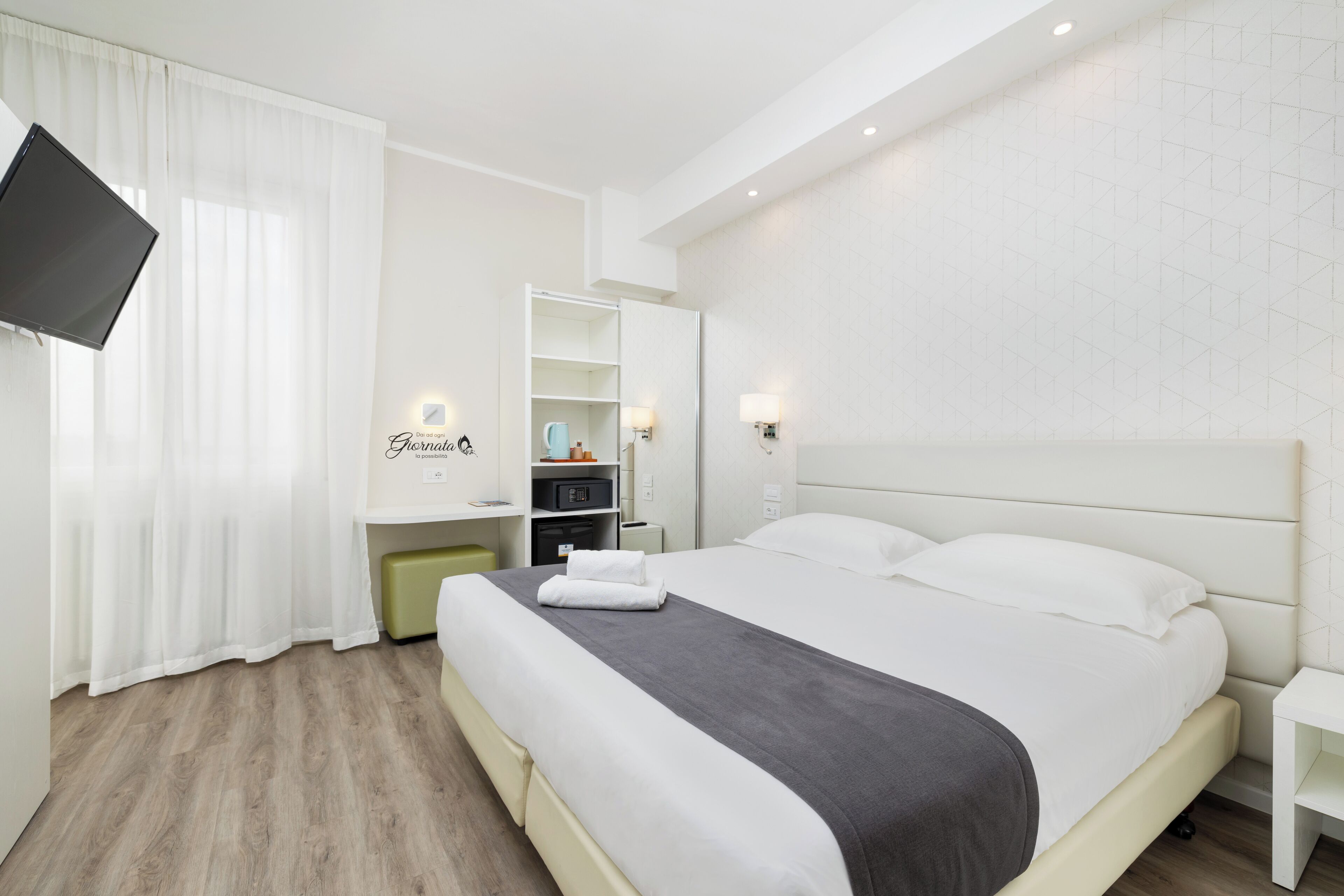 A modern hotel room with a large bed, a flat-screen TV mounted on the wall, a bedside table, and a wardrobe.
