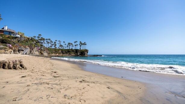 Una playa arenosa con huellas, cielo azul despejado y una casa con palmeras sobre un acantilado.